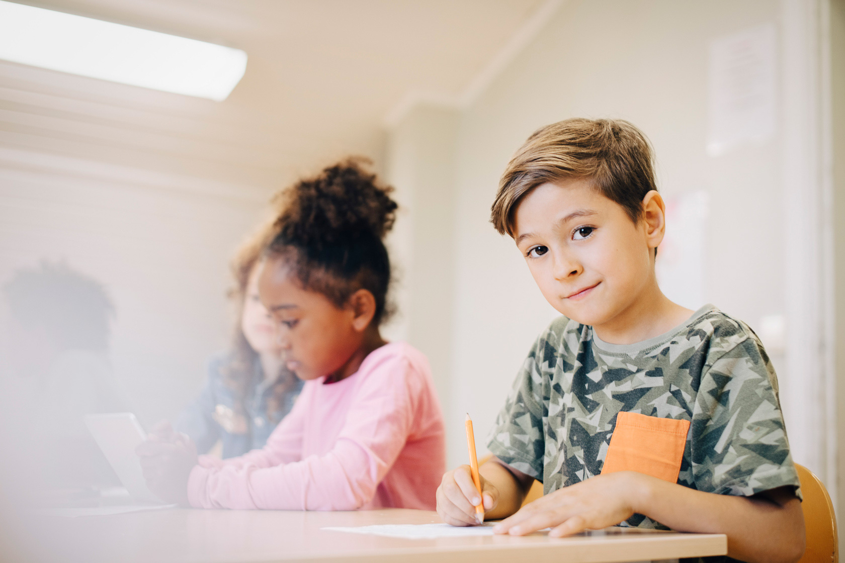 Dekorationsbild Viljaskolan. En pojke sitter vid sitt skrivbord i en skola. Han arbetar med papper och penna och ser nöjd ut när han tittar mot kameran. Några klasskamrater syns i bakgrunden.