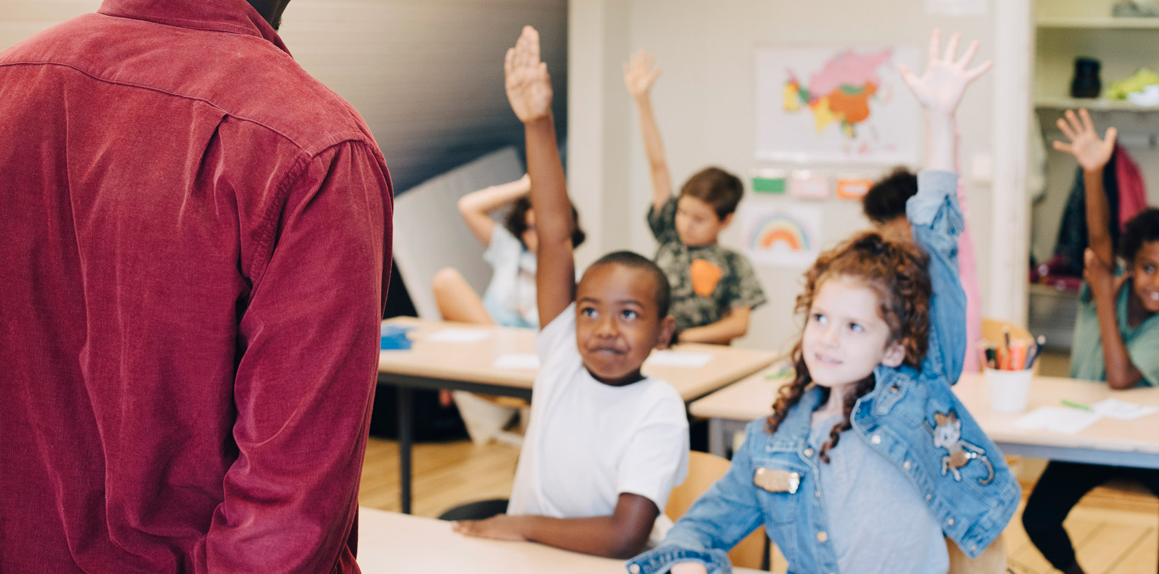 Dekorationsbild Viljaskolan. Elever i ett klassrum i en grundskola räcker upp sina händer för att svara på en fråga. En lärare står framför dem.